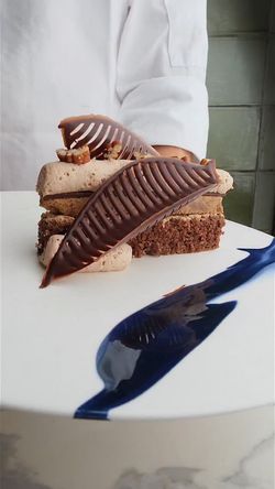 Chocolate dessert with textured chocolate leaves and nut garnish on a white plate with blue sauce streak.