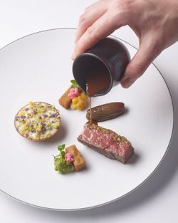 Hand pouring sauce over a delicately plated piece of steak with colorful vegetables and garnishes on a white textured plate.