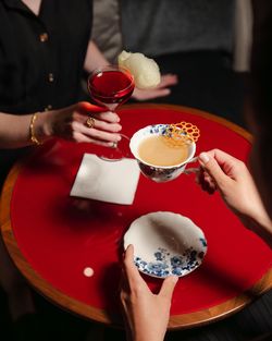Two people on a red round table hold a red cocktail and a decorated tea cup with a matching saucer.