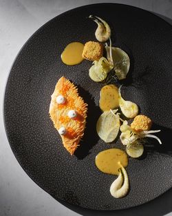 Fine dining plating with creamy sauces, vegetable garnishes, and breaded elements on a dark textured plate.