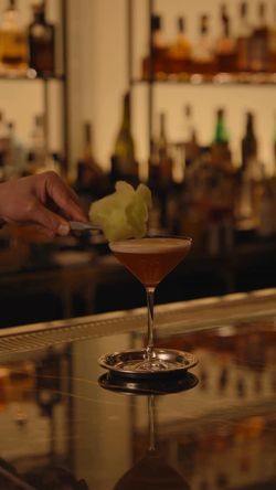 A bartender garnishes a cocktail with a green foam or garnish at a dimly lit upscale restaurant bar.