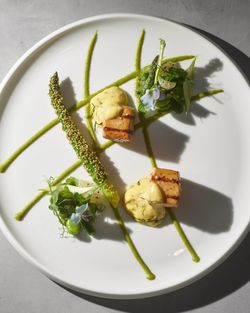 Grilled fish cubes with yellow sauce, sesame-crusted asparagus, greens, edible flowers, and green sauce lines on a plate.