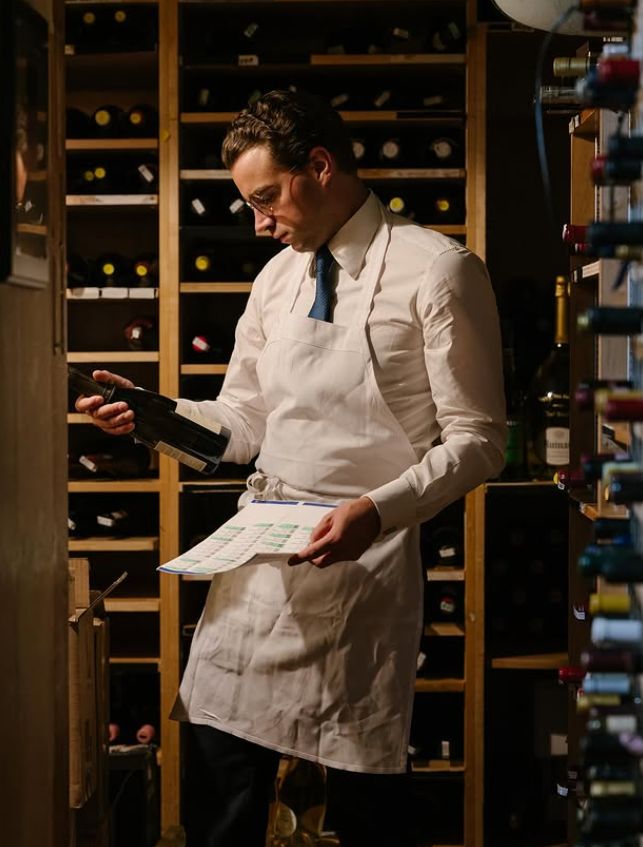 A sommelier holding a wine bottle and a menu in a wine cellar filled with various bottles.